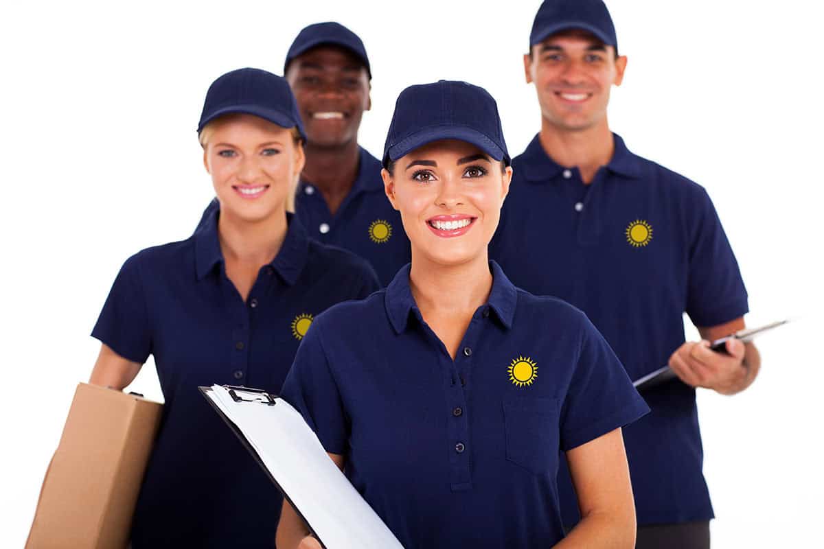 A group of four smiling individuals in new New Braunfels custom apparel in navy blue polo shirts and caps, two holding clipboards and one carrying a box, standing together against a white background.